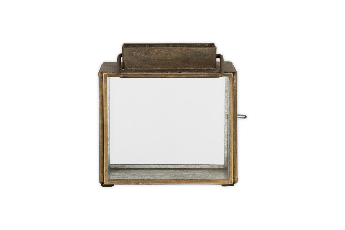 The Obi Mini Table Lantern by nkuku features an antique brass square frame, glass sides, a rectangular handle on top, and a glass door for easy access, shown against a plain white background.