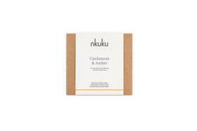 A rectangular box with brown sides and a white label reading “nkuku Cardamom & Amber 3 Wick Scented Candle,” a natural wax blend with sandalwood notes in a recyclable glass holder.