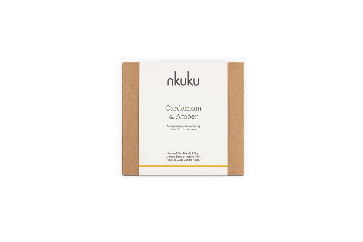 A rectangular box with brown sides and a white label reading “nkuku Cardamom & Amber 3 Wick Scented Candle,” a natural wax blend with sandalwood notes in a recyclable glass holder.