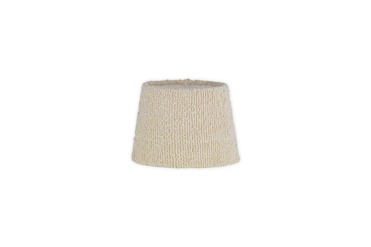 The Sakiti Bouclé Lampshade by nkuku features an off white, textured design with a gently tapered cylindrical shape. Crafted from cozy bouclé material, it adds ambient lighting to any space. The product image has a plain white background.