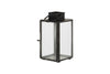 The nkuku Sia Mini Table Lantern - Black features clear glass panels, a top handle, and a hinged door for a tealight. Its rectangular metal frame stands on four small feet, adding charm to any setting.