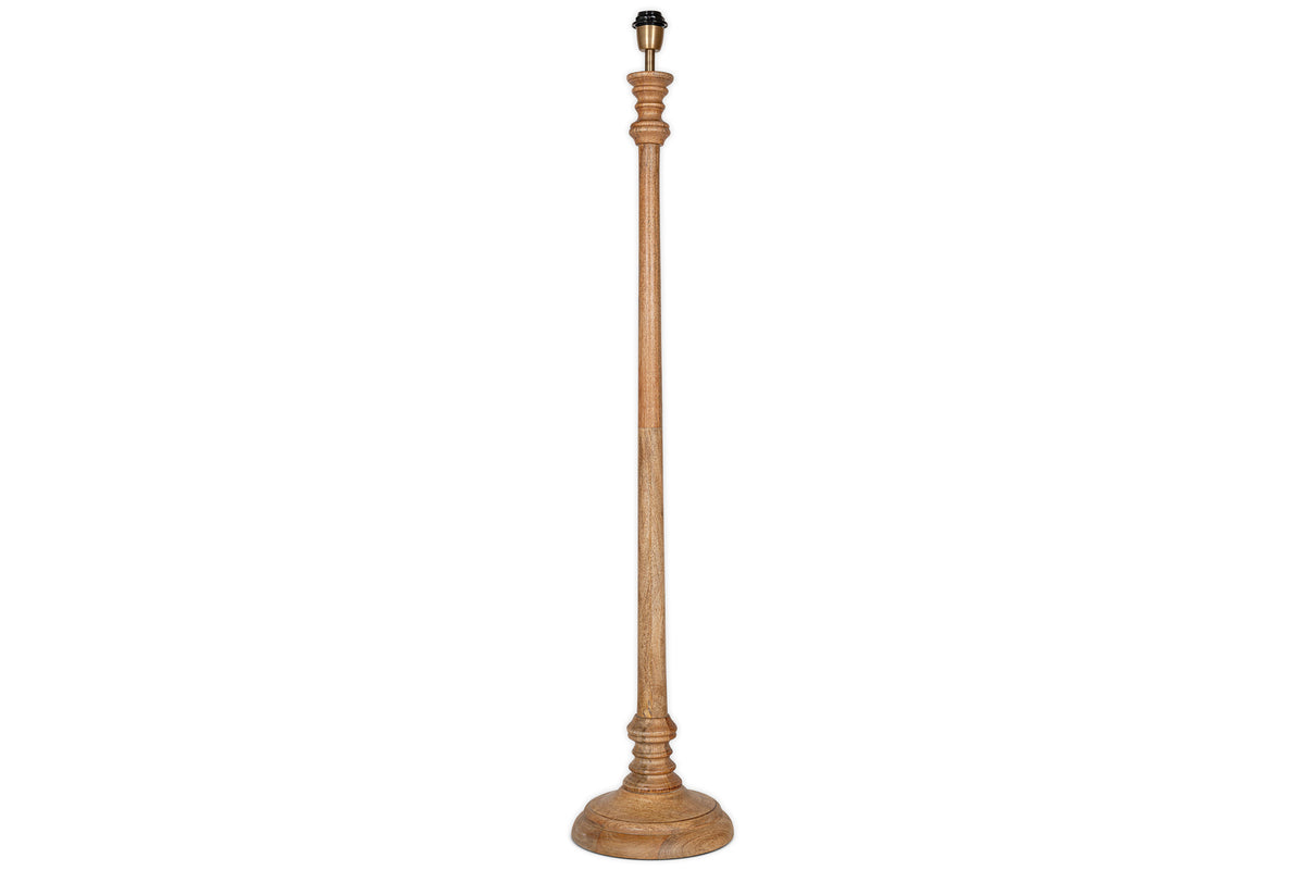 The Usha Classic Wooden Floor Lamp - Natural by nkuku is a tall, handmade mango wood base with a natural finish, round sturdy bottom, and ornately carved slender pole. Eco-friendly, it comes without a lampshade or bulb. Background is white.