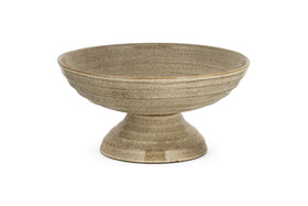 The Valinha Earthenware Footed Bowl by nkuku features a wide, shallow grey dish with a flared base and reactive glaze, ideal for enhancing rustic tablescapes. Displayed on a white background.