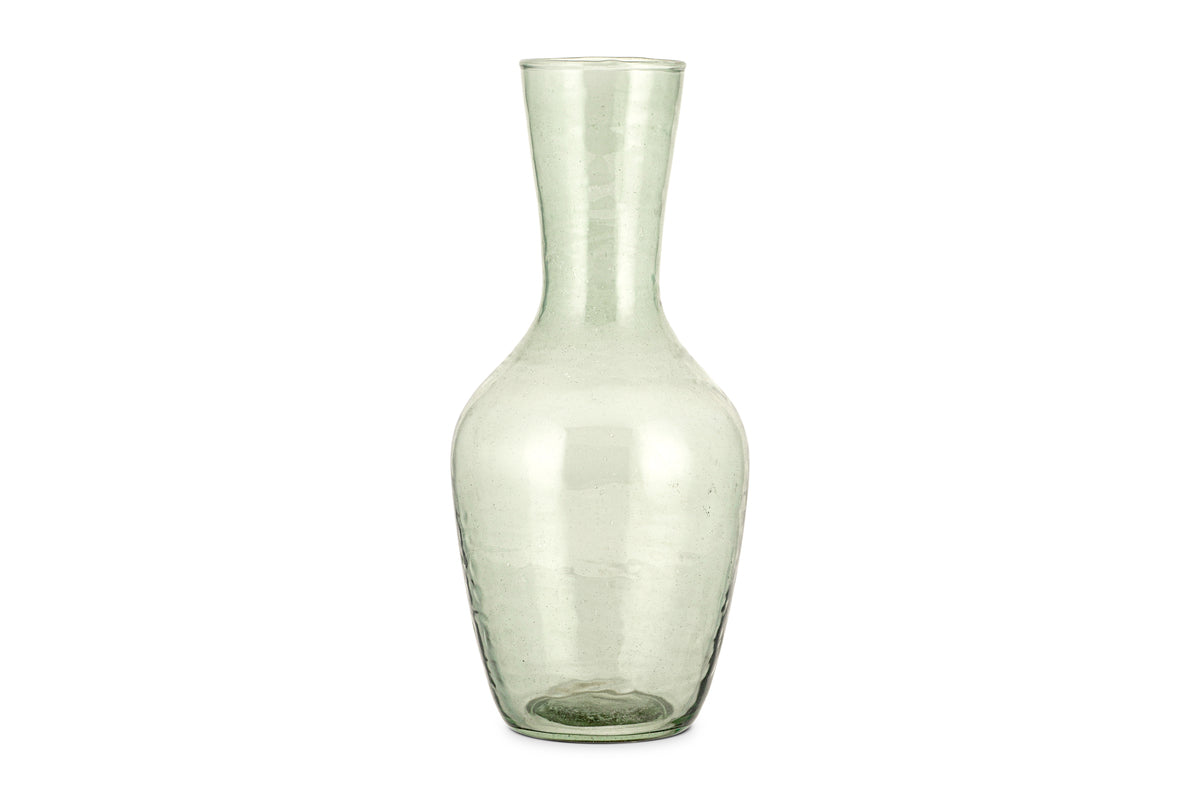 The Yala Hammered Jug - Sage Green by nkuku is a tall, clear glass jug with a narrow neck and wider base, crafted from subtly textured, slightly green-tinted recycled glassware.