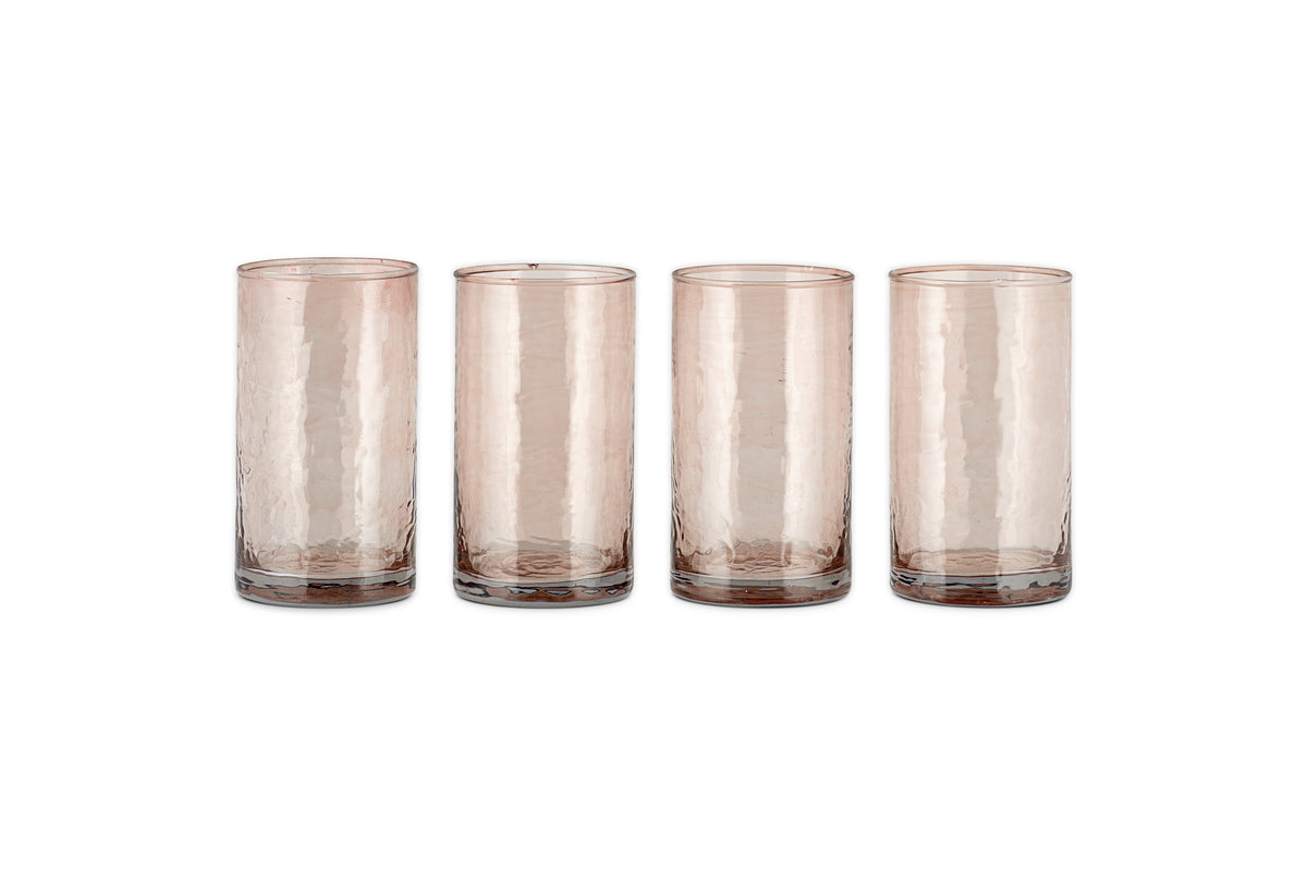 A set of four Yala Hammered Highball glasses by nkuku, made from recycled glass in a dusty terracotta hue, are arranged in a row on a white background.