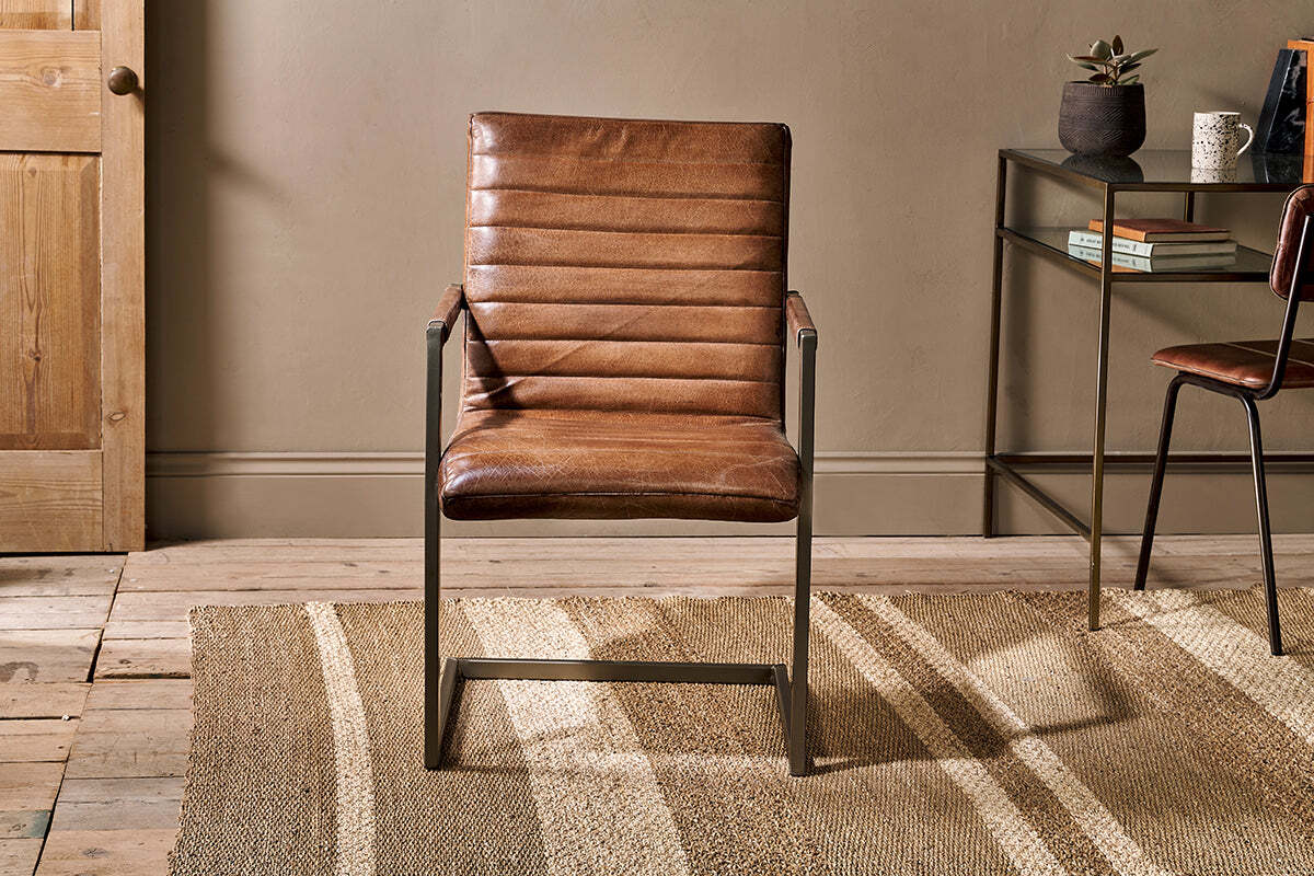 The Wamma Leather Desk Chair by nkuku, featuring distressed dark brown leather and a ribbed design, sits on a jute rug in front of a tan wall.