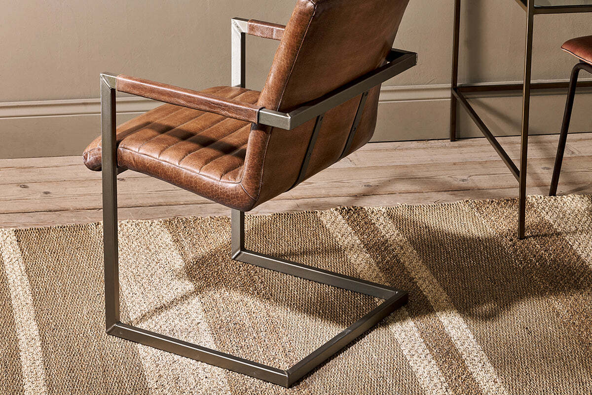 A close-up of the nkuku Wamma Leather Desk Chair, featuring dark brown ribbed leather and a metal frame, sits on a woven rug over a wooden floor.