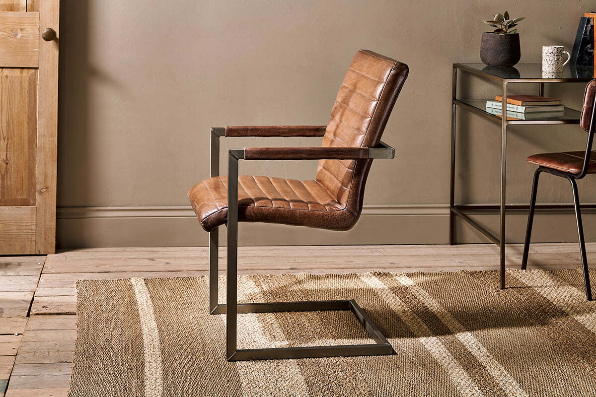 The Wamma Leather Desk Chair by nkuku, featuring quilted dark brown leather and a black metal cantilever frame, sits on a beige striped rug in a room with wooden floors, neutral walls, a wooden door, and a decor-adorned side table.