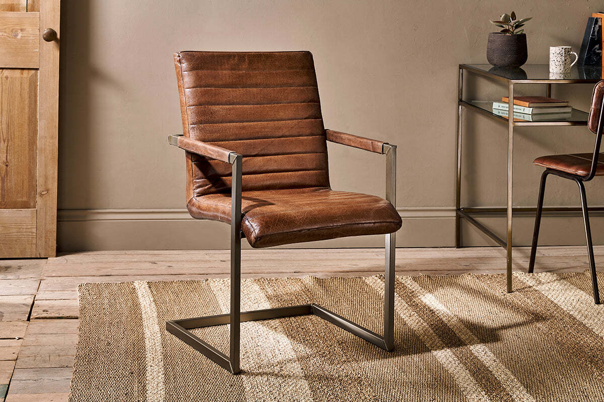 The Wamma Leather Desk Chair by nkuku, featuring dark brown leather with a ribbed design and a cantilever metal frame, sits on a beige textured rug in a cozy room with wood flooring, tan walls, and a glass console table in the background.