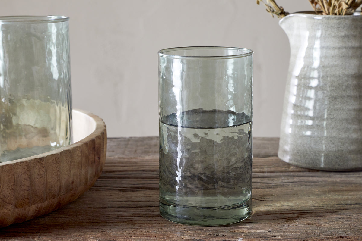 The nkuku Yala Hammered Highball - Sage Green (Set of 4) is shown half-filled with water on a rustic wooden table, beside a wooden bowl and a ceramic jug holding dried flowers.