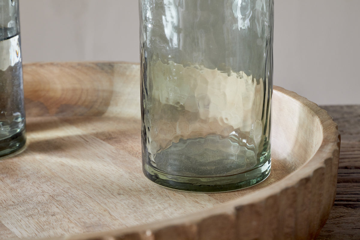 A set of four nkuku Yala Hammered Highball glasses in Sage Green sits on a round, light-finished wooden tray atop a wood surface, with part of another cocktail glass visible to the left.