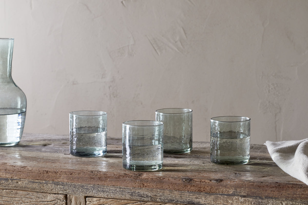 Four Yala Hammered Tumblers in sage green by nkuku, filled with water, rest on a rustic wooden table. A partial glass carafe sits to the left and a white cloth is draped on the right, with a plain textured wall in the background.