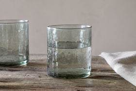 A sage green, hammered recycled glass tumbler from the Yala Hammered Tumbler - Sage Green (Set of 4) by nkuku is placed on a rustic wooden surface, with part of another tumbler and a folded white cloth nearby against a neutral background.