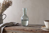A clear recycled Yala glassware carafe half-filled with water sits on a rustic wooden table next to stacked beige bowls, the nkuku Yala Hammered Jug in Sage Green filled with dried flowers, and a folded linen cloth.