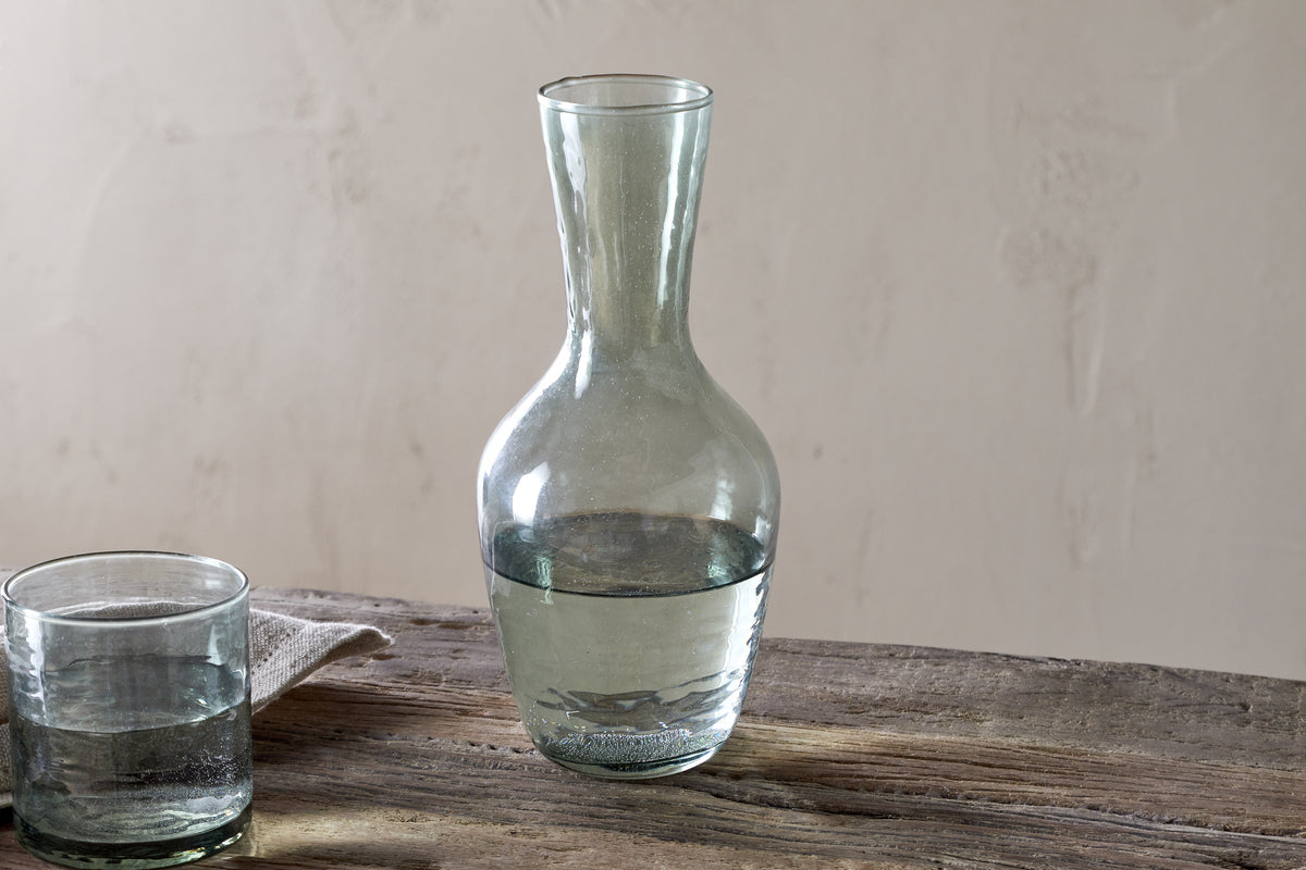 The nkuku Yala Hammered Jug in sage green, partially filled with water, sits on a rustic wooden table beside a glass tumbler and folded cloth, set against a neutral background.