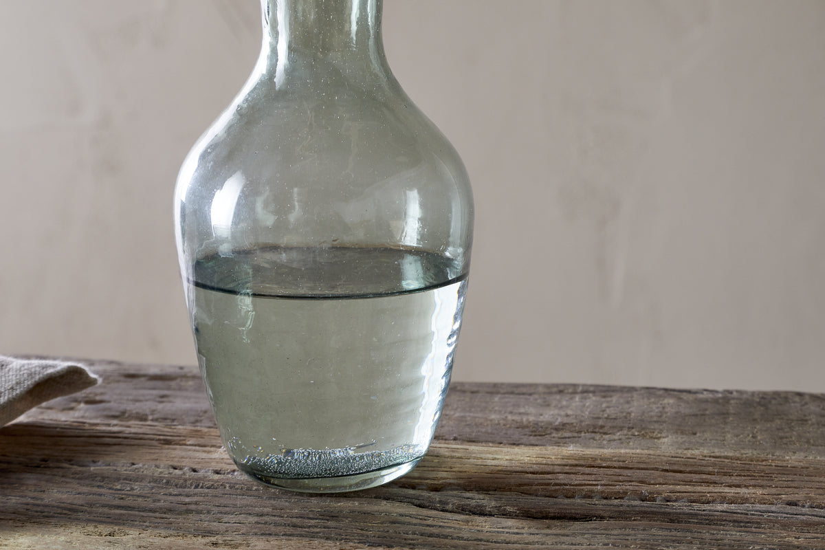 A nkuku Yala Hammered Jug in sage green, filled halfway with water, sits on a rustic wooden surface with a neutral beige background and a light-colored cloth partly visible to the left.