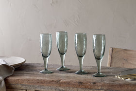 A set of four nkuku Yala Hammered Champagne Glasses in Sage Green is displayed on a rustic wooden table with plates, cutlery, and a napkin nearby, set against a light beige textured wall.