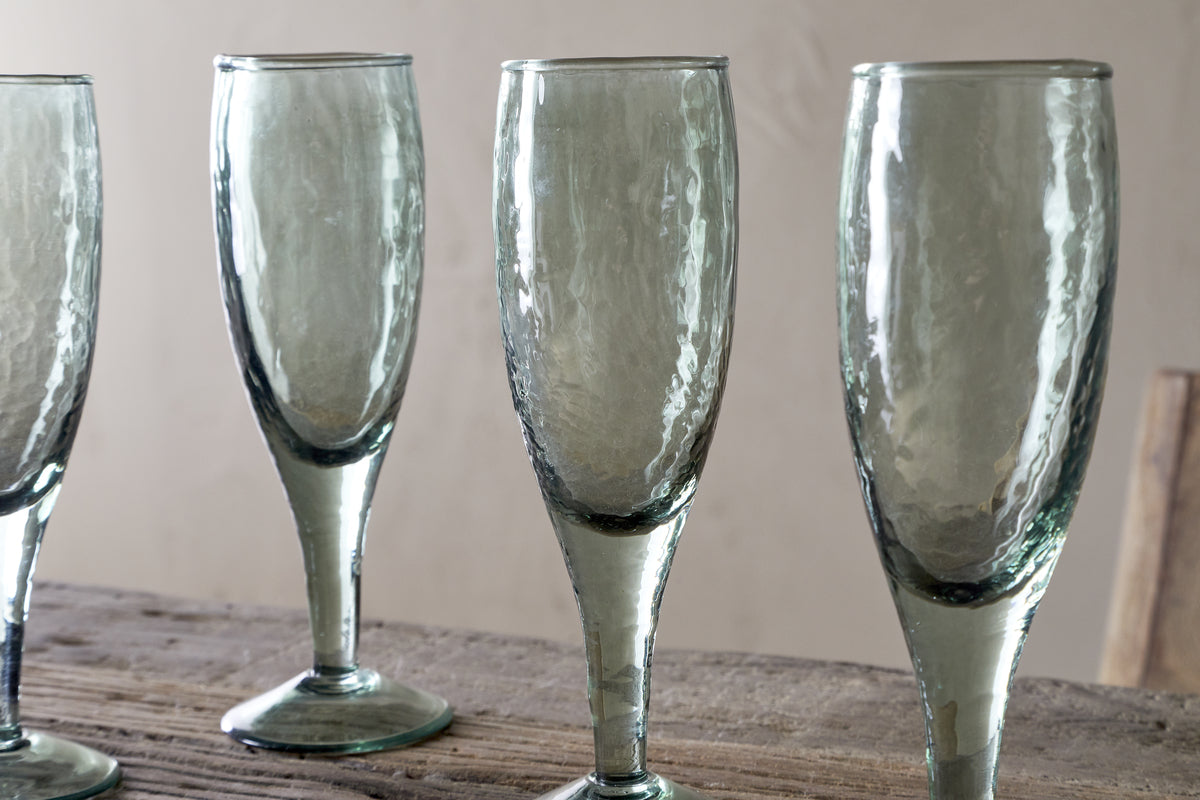 Four Yala Hammered Champagne Glasses in sage green by nkuku stand on a rustic wooden surface against a beige background. The set of four glasses is partially cropped, showing three fully and part of the fourth.