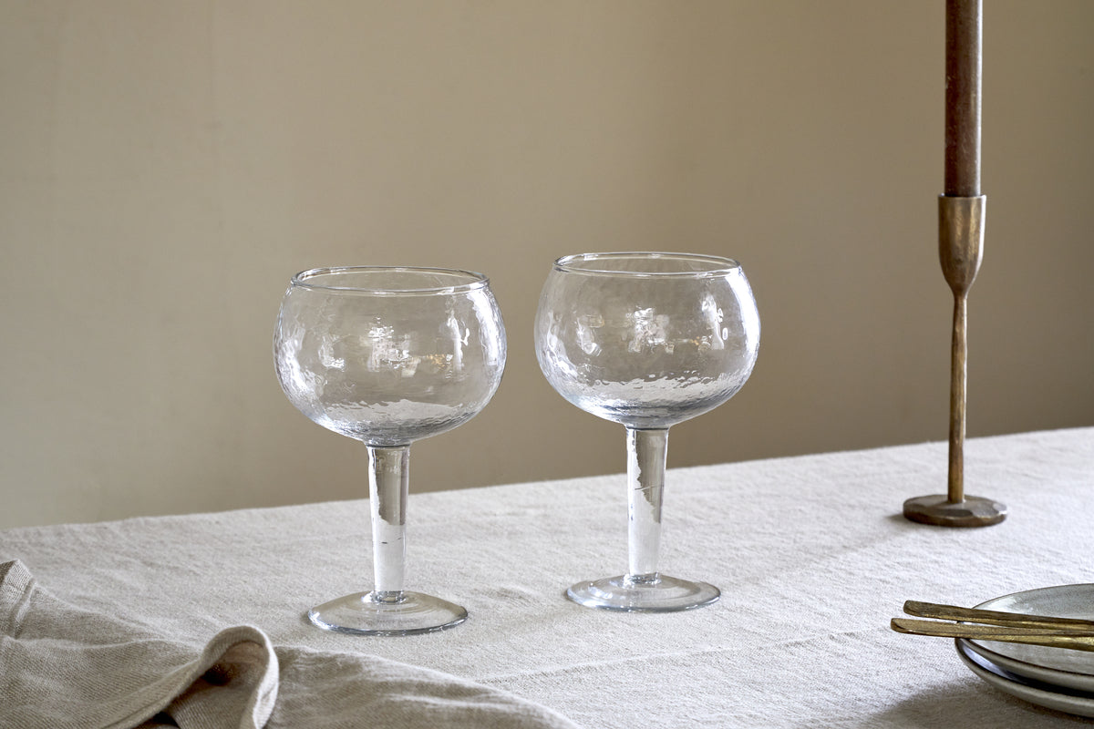 Two nkuku Yala Hammered Gin Glasses (Set of 2) in clear, with textured design, rest on a light tablecloth next to a brass candlestick, gold utensils, and a folded napkin against a plain beige wall.