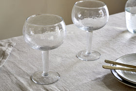 A set of two nkuku Yala Hammered Gin Glasses in clear, featuring a textured finish, rests on a beige tablecloth beside stacked plates with gold-toned cutlery and a glass of water in the background.