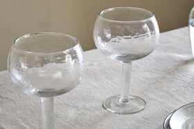 Two empty Yala Hammered Gin Glasses - Clear (Set of 2) by nkuku, with elegant stems and a hammered texture, rest on a white textured tablecloth. The softly lit neutral background highlights their refined design.