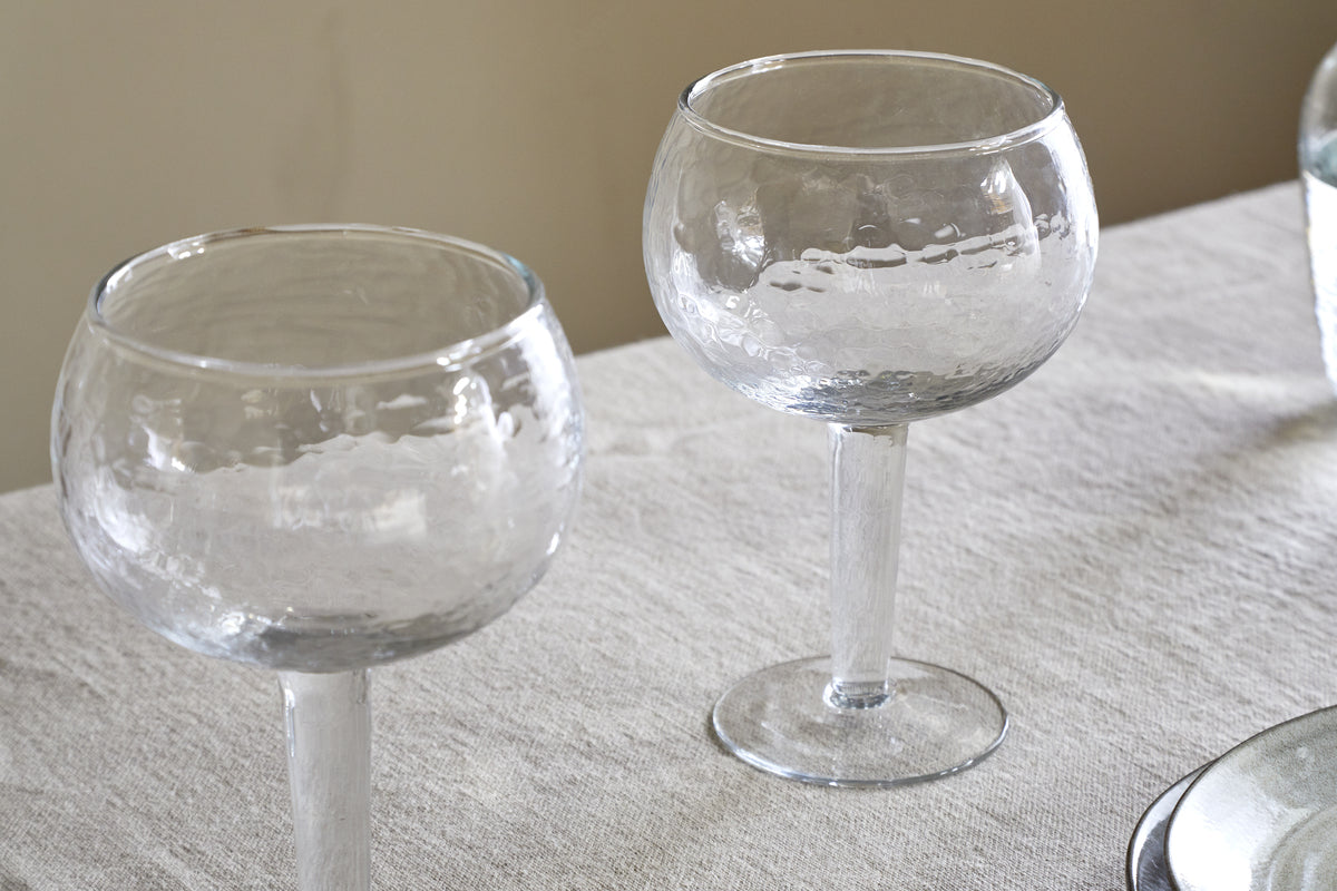 Two empty Yala Hammered Gin Glasses - Clear (Set of 2) by nkuku, with elegant stems and a hammered texture, rest on a white textured tablecloth. The softly lit neutral background highlights their refined design.