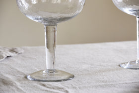 Close-up of the base and stem of two nkuku Yala Hammered Gin Glasses (Clear, Set of 2) on a light-textured tablecloth; a third goblet is partially visible on the right.
