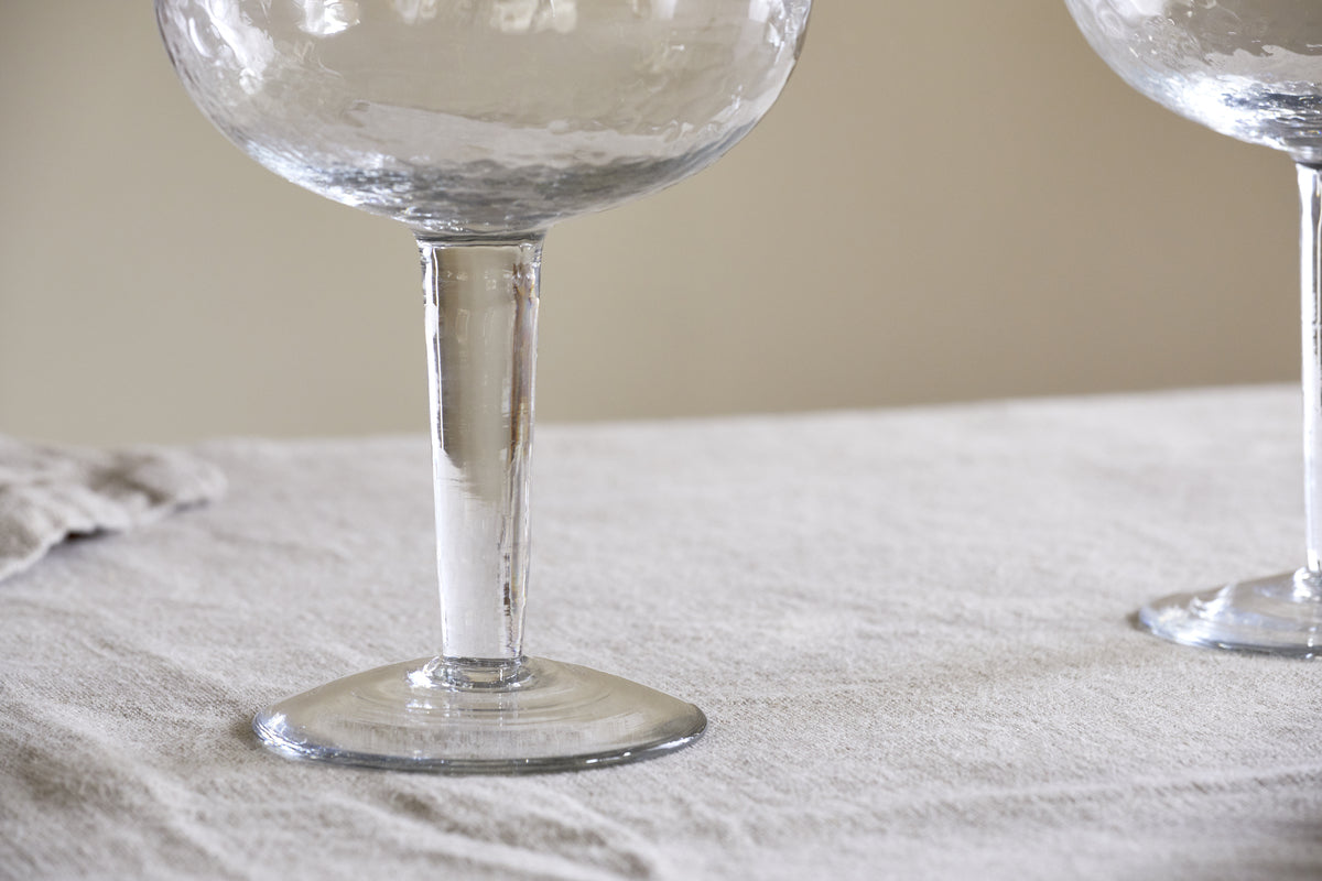 Close-up of the base and stem of two nkuku Yala Hammered Gin Glasses (Clear, Set of 2) on a light-textured tablecloth; a third goblet is partially visible on the right.