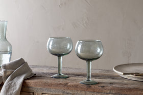 The nkuku Yala Hammered Gin Glass - Sage Green (Set of 2) features two round, clear glasses with green stems on a rustic wooden table, alongside a folded beige cloth, glass carafe, and plate against a textured neutral wall.