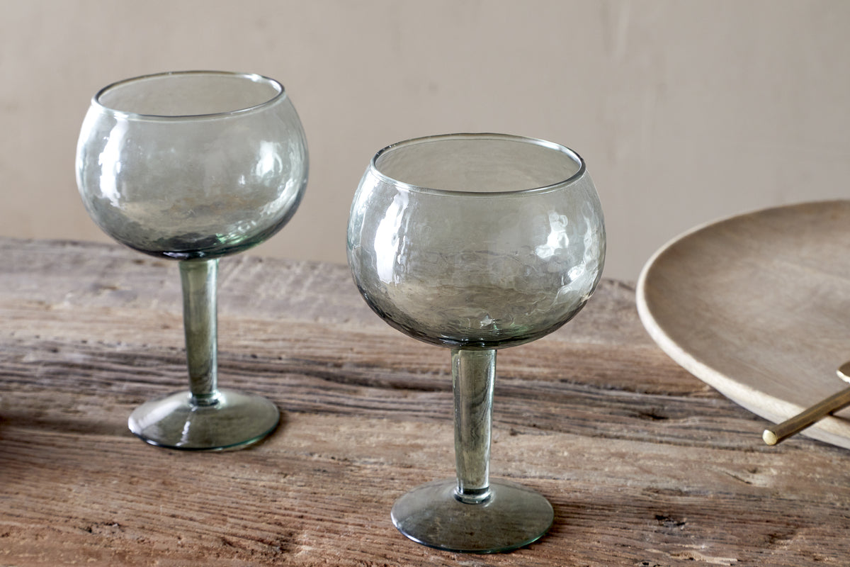 Two Yala Hammered Gin Glasses in sage green by nkuku, featuring short stems and a unique texture, are displayed on a rustic wooden table next to a beige plate.