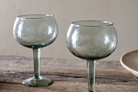 The nkuku Yala Hammered Gin Glasses in sage green, sold as a set of 2, feature round hammered glass and short stems. They sit empty on a rustic wooden surface with a neutral background and a beige plate partially visible.