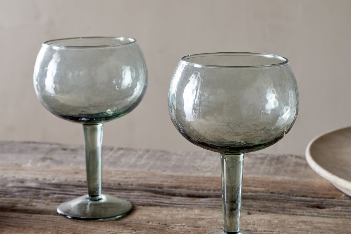 The nkuku Yala Hammered Gin Glasses in sage green, sold as a set of 2, feature round hammered glass and short stems. They sit empty on a rustic wooden surface with a neutral background and a beige plate partially visible.