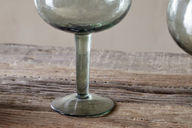 A close-up of the base and stem of an nkuku Yala Hammered Gin Glass in sage green, from a set of 2, displayed on a rustic wooden surface.