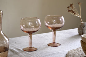 Two nkuku Yala Hammered Gin Glasses in Dusty Terracotta sit on a light tablecloth next to the matching pitcher and neutral ceramic bowls, with dried flowers in the background.