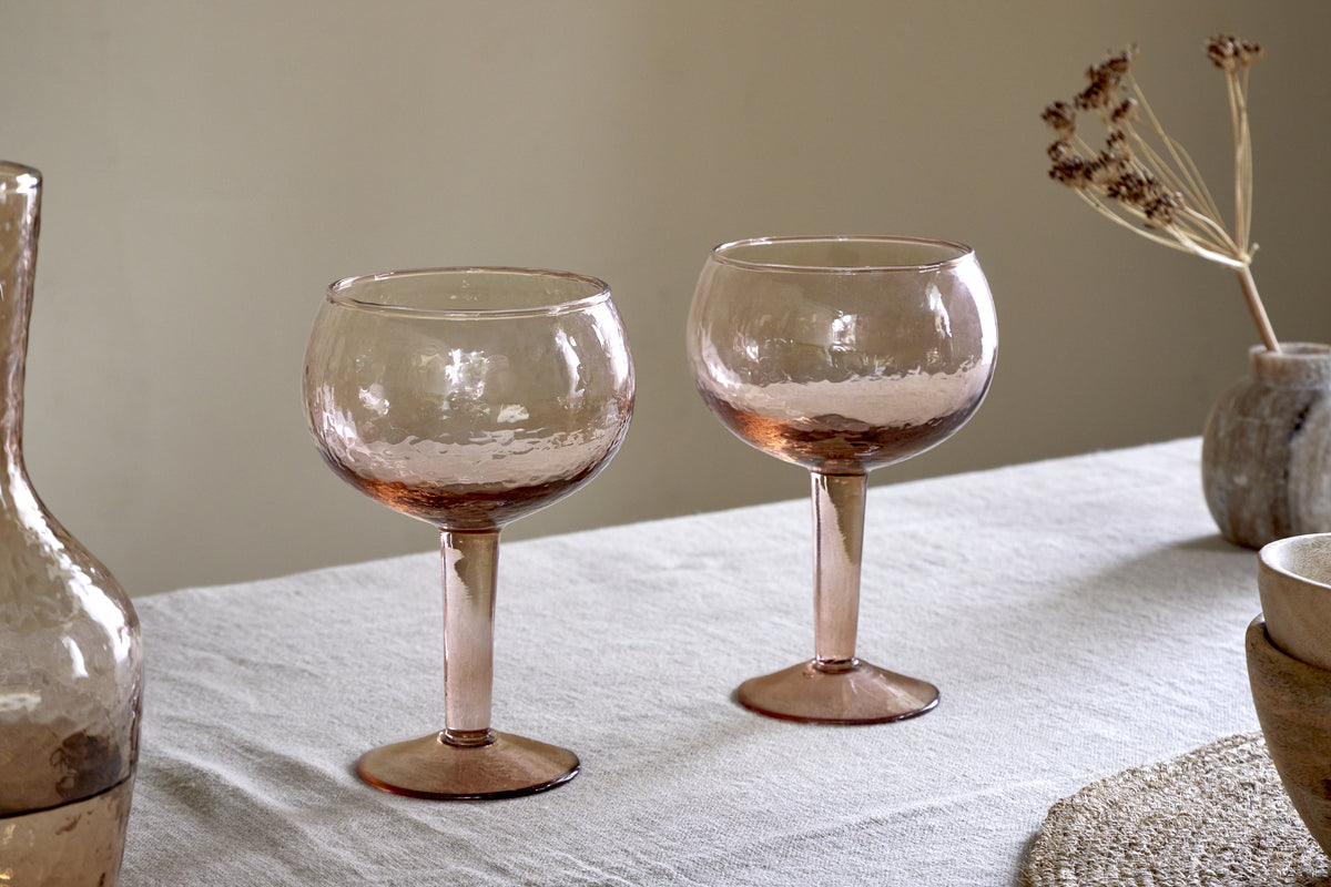 Two nkuku Yala Hammered Gin Glasses in Dusty Terracotta sit on a light tablecloth next to the matching pitcher and neutral ceramic bowls, with dried flowers in the background.