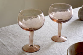 A set of two nkuku Yala Hammered Gin Glasses in Dusty Terracotta rests on a light fabric tablecloth, soft lighting highlighting the textured glass and warm pink tones.