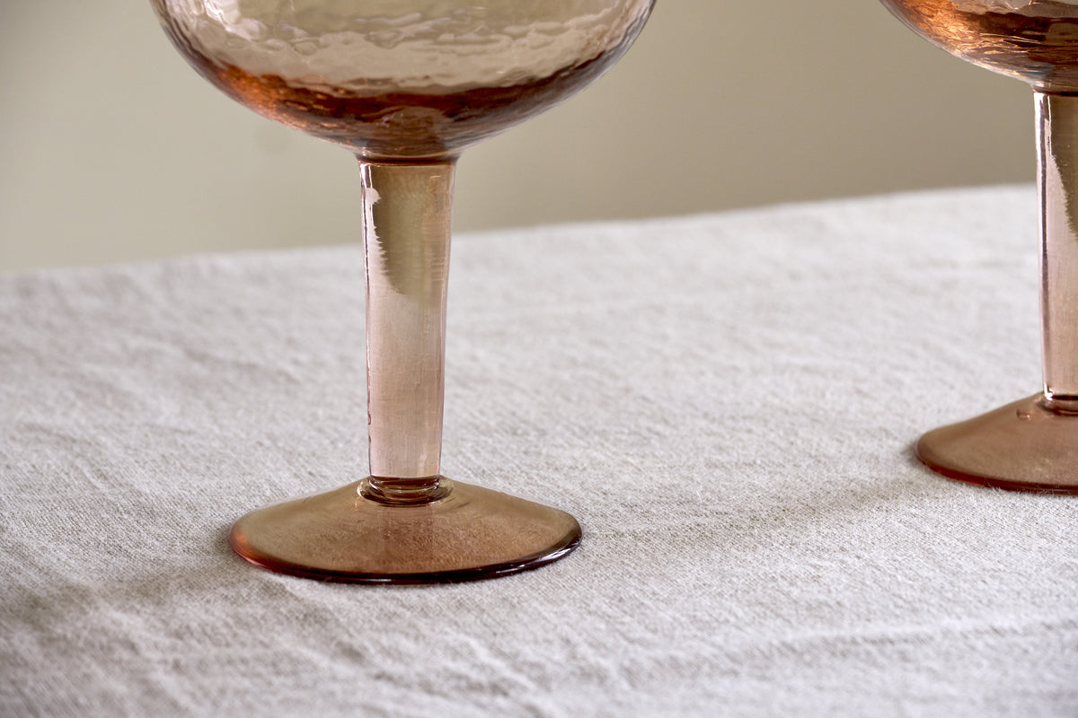 Two Yala Hammered Gin Glasses in Dusty Terracotta by nkuku sit on a light, textured fabric surface, showing only the hammered lower bowls.
