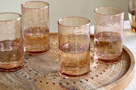 A set of four nkuku Yala Hammered Highball glasses in Dusty Terracotta sits on an ornately carved wooden tray with geometric patterns, displayed on a light-colored surface.