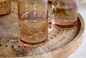 Three nkuku Yala Hammered Highball glasses in Dusty Terracotta, filled with a light brown liquid, sit on a round carved wooden tray with geometric patterns atop a light-colored tablecloth. Set of 4.