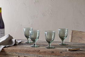A set of four nkuku Yala Hammered Wine Glasses in Sage Green is arranged on a rustic wooden table with a folded linen napkin, corkscrew, and a partially seen wine bottle against a plain, light-colored wall.