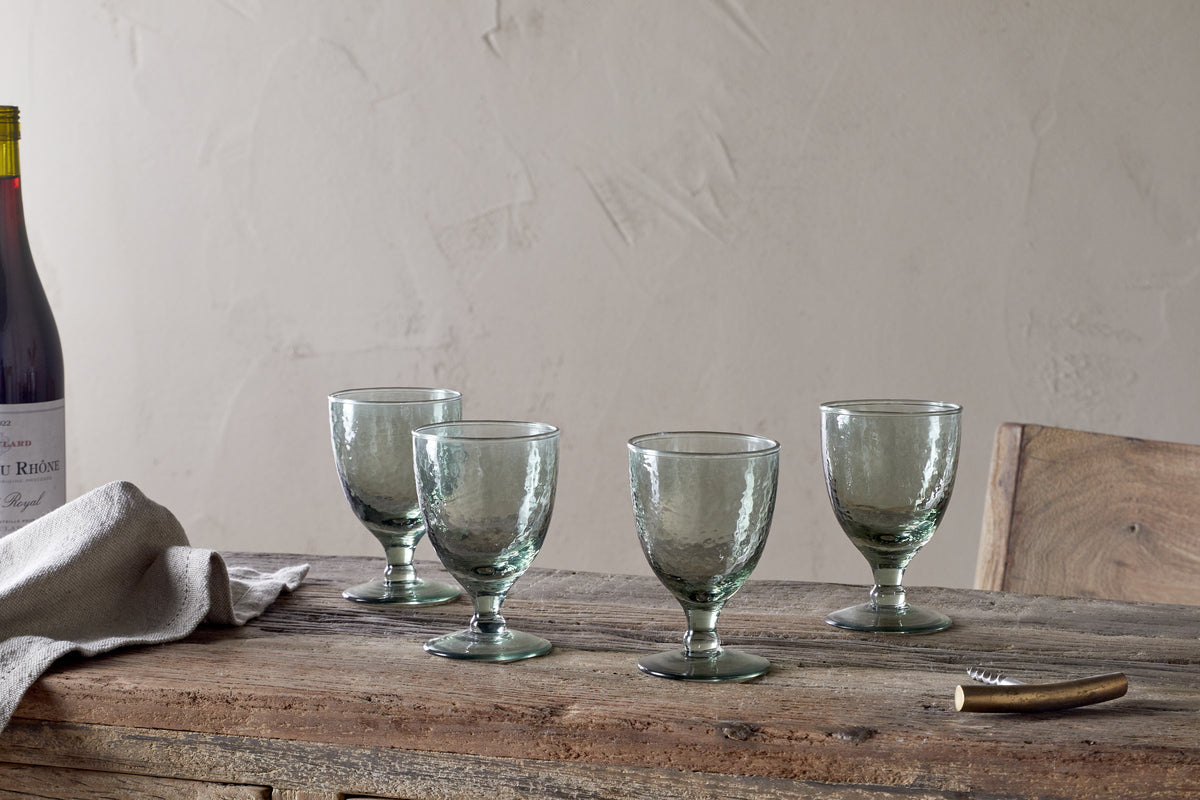 A set of four nkuku Yala Hammered Wine Glasses in Sage Green is arranged on a rustic wooden table with a folded linen napkin, corkscrew, and a partially seen wine bottle against a plain, light-colored wall.