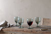 Four nkuku Yala Hammered Wine Glasses in sage green are on a rustic wooden table; three are empty, one holds red wine. A beige napkin is folded on the left, and a small knife is placed to the right.
