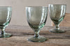 Four Yala Hammered Wine Glasses in sage green by nkuku, each with a short stem, are placed on a rustic wooden table against a softly blurred, neutral background.