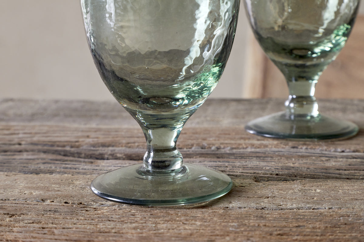Close-up of the nkuku Yala Hammered Wine Glass in Sage Green (Set of 4), featuring thick stems on a rustic wooden surface, with one glass in sharp focus and another softly blurred in the background.