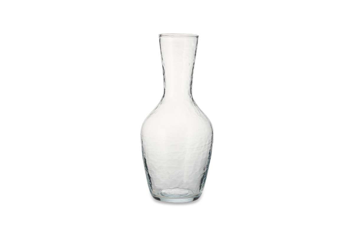 The nkuku Yala Hammered Jug - Clear Glass is displayed against a plain white background. Featuring a narrow neck, flared base, and subtly hammered finish, the empty jug casts a faint shadow.