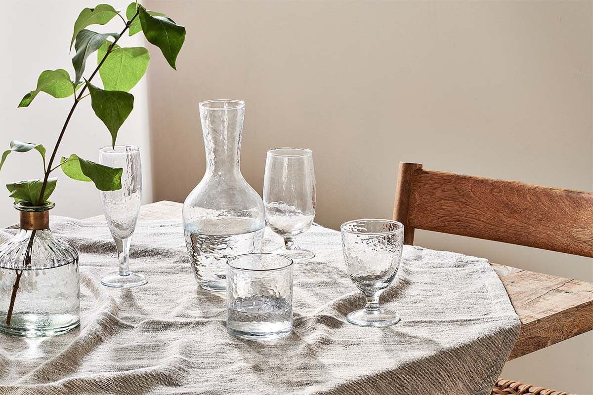 A wooden table with a light striped cloth holds a clear glass vase with green leaves, a carafe, and nkuku's Yala Hammered Tumblers in clear glass (set of 4), creating an organic look. A wooden chair with woven seat sits nearby in the soft sunlight.