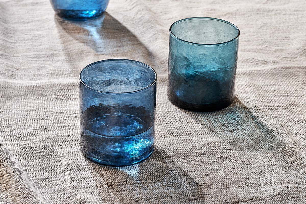 Two nkuku Yala Hammered Tumblers in indigo from the set of 4 rest on a light beige, textured fabric. Sunlight casts soft shadows and highlights, bringing out the glasses’ transparency and the woven look of the surface below.