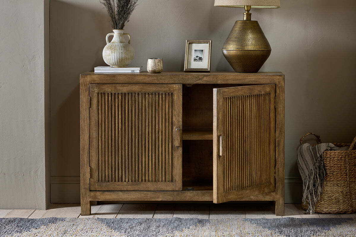 The nkuku Yasha Reeded Mango Wood Sideboard with ribbed doors, one ajar, stands by a beige wall. On top: lamp, framed photo, vase of dried flowers, candle, and book. A woven basket is beside it on a wooden floor with a patterned rug.
