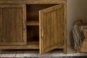 The nkuku Yasha Reeded Mango Wood Sideboard features fluted doors, one slightly open to reveal shelves inside. To the right sits a woven basket with a striped throw.