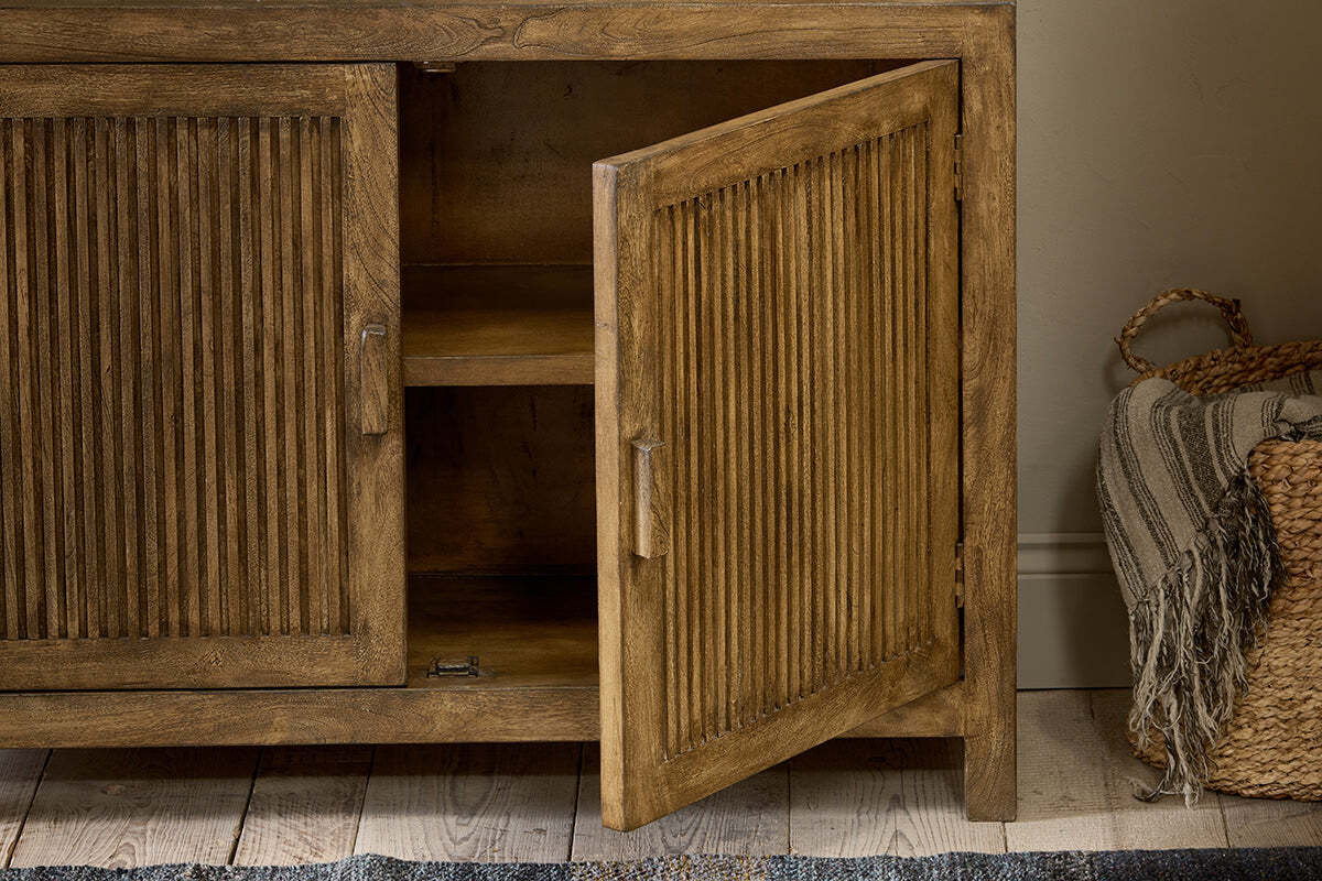 The nkuku Yasha Reeded Mango Wood Sideboard features fluted doors, one slightly open to reveal shelves inside. To the right sits a woven basket with a striped throw.